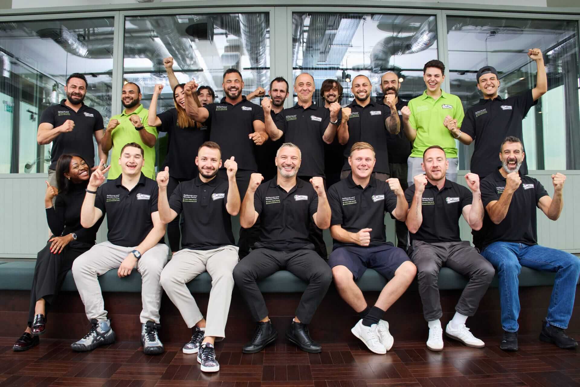 The enthusiastic Mr. Sander® team poses together, wearing black and green branded shirts, showing their excitement and professionalism. The team is gathered in front of large windows, sitting on a bench, and cheering. The floor beneath them is a dark, polished hardwood, reflecting their expertise in floor refinishing.