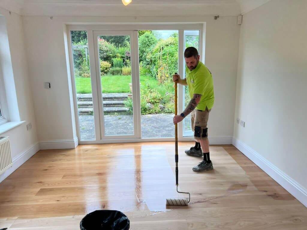 A professional from Mr Sander® applying a lacquer finish to a freshly sanded wooden floor in a bright room with large windows overlooking a lush garden. The worker is using a roller to ensure an even application, enhancing the floor's natural beauty.