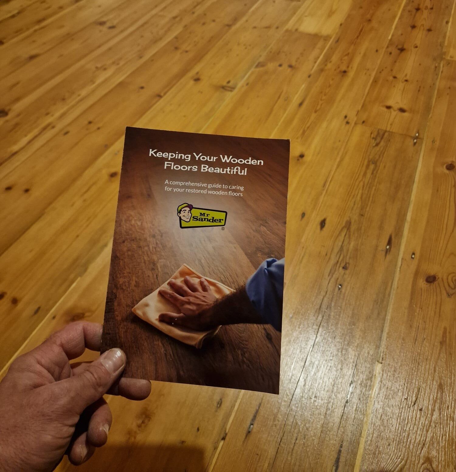 Hand holding a ‘Keeping Your Wooden Floors Beautiful’ leaflet from Mr Sander® above a freshly restored pine floor – illustrating expert floor restoration results and after-care guidance.