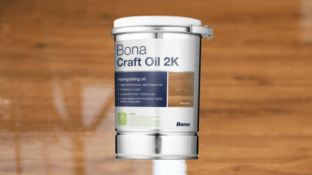 A silver container of Bona Craft Oil 2K is displayed on a wooden floor. The label describes it as a high-performance, plant-based impregnating oil that provides protection in one coat. It offers a long open time, quick cure, and can be overcoated with the Bona Traffic family of lacquers. The product is shown in a 1.25L size.