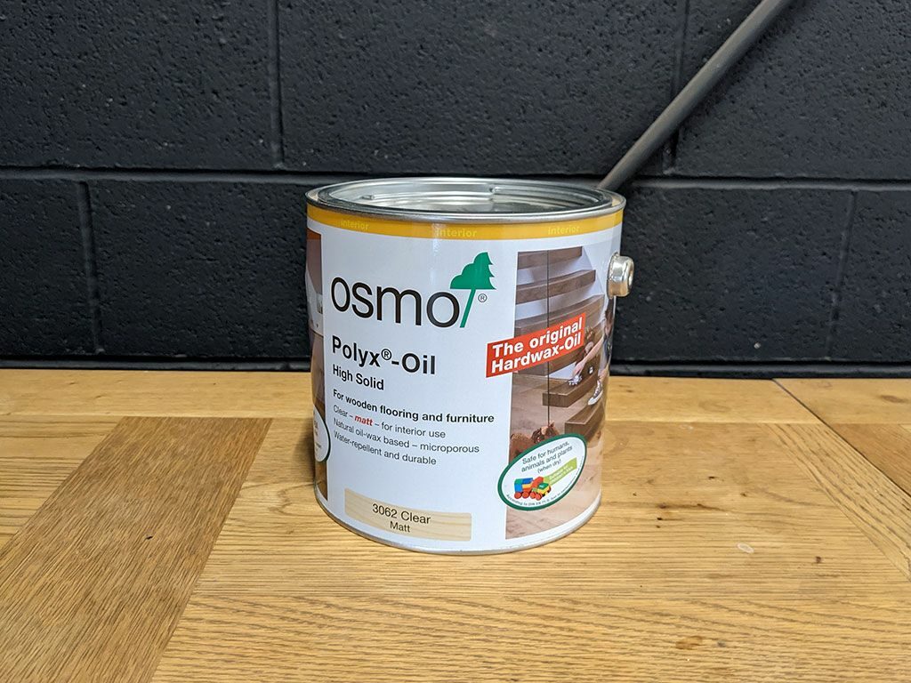 A can of Osmo Polyx®-Oil, labeled "The original Hardwax-Oil," on a wooden surface against a black brick wall.