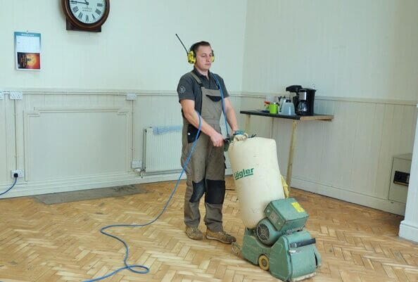 A professional floor sanding technician in the UK operating a Lagler belt sander on a herringbone parquet floor, part of an Overnight Floor Sanding project.