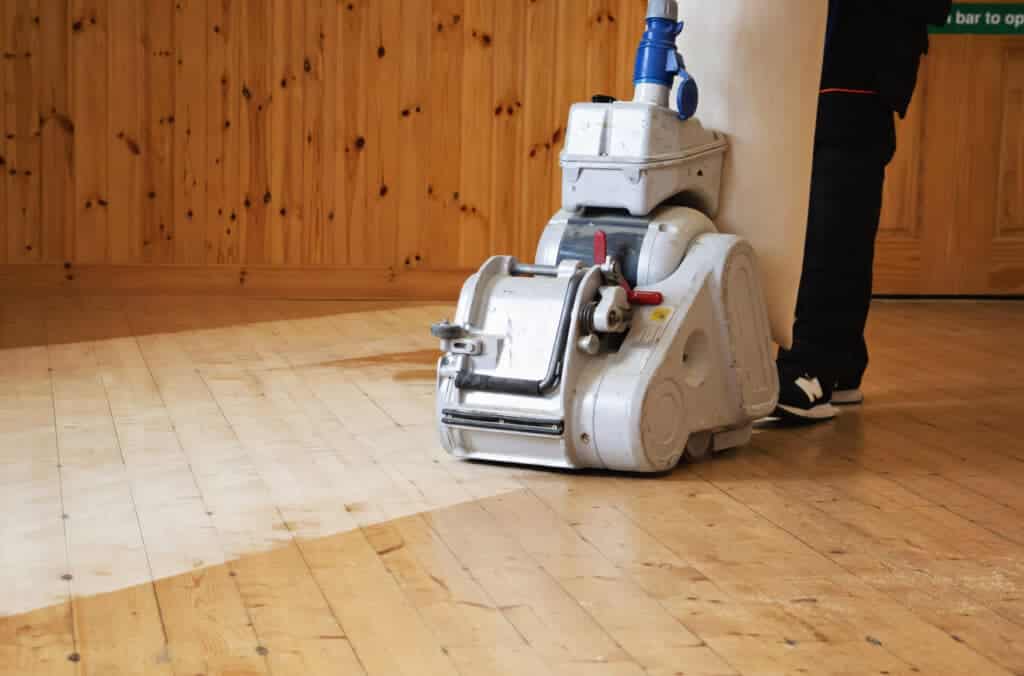 A professional from Mr Sander® sanding a pine floor in a living room using the Bona Belt UX (AMK173100) machine. The Bona Belt UX features a 2.2 kW motor, 230 V voltage, 50 Hz frequency, 200 mm drum width, and 200x750 mm sanding belt size, weighing 85 kg.