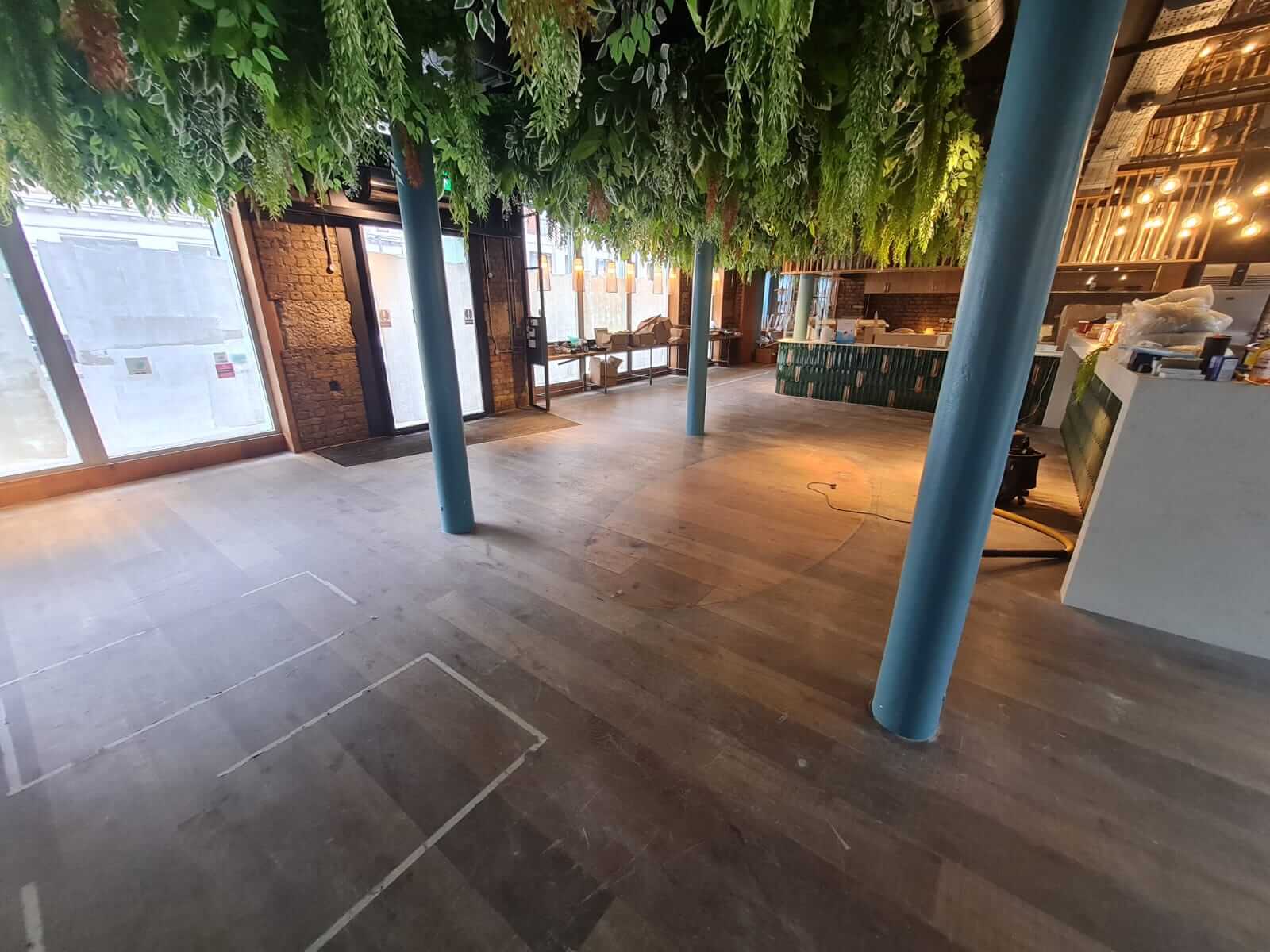 A spacious room with wooden floors in the process of being sanded and refinished by Mr Sander®. The floor shows signs of ongoing work, with some areas more polished than others. The room features blue columns, green hanging plants from the ceiling, large windows, and a bar area with green tiles. Exposed brick walls and various tools and materials are visible, indicating renovation activity.