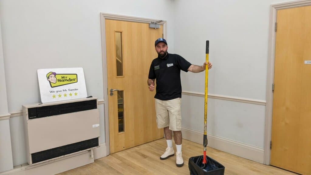  A Mr Sander® technician stands in a room with freshly sanded and finished wooden floors. He is dressed in a black shirt, beige shorts, white socks, and a blue cap. The technician holds a mop with a yellow handle, and a mop bucket is nearby. A Mr Sander® sign with a five-star rating is displayed on a wall-mounted heater.