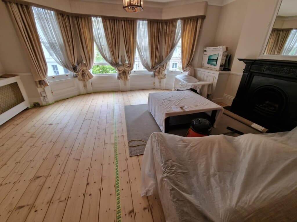 A spacious living room with large bay windows and light curtains, showcasing freshly sanded pine wood floors. The furniture is covered with protective sheets, and a vacuum cleaner is visible on the side. The room features neutral beige walls, a black decorative fireplace, and a chandelier hanging from the ceiling.