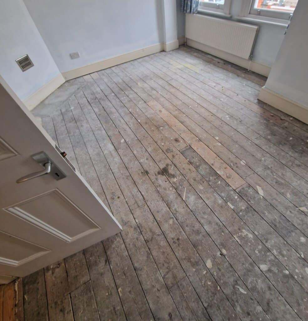 A room with natural wood plank flooring that appears weathered and covered with marks and stains. The floorboards are in need of restoration, with a visible patch near the center where some planks have been replaced. The room has white walls, a radiator under the window, and baseboards painted in a light cream color.