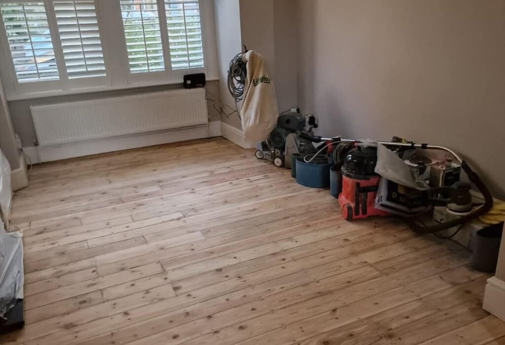 A partially sanded pine wooden floor with sanding equipment neatly placed along the wall in a bright room with large windows and shutters.