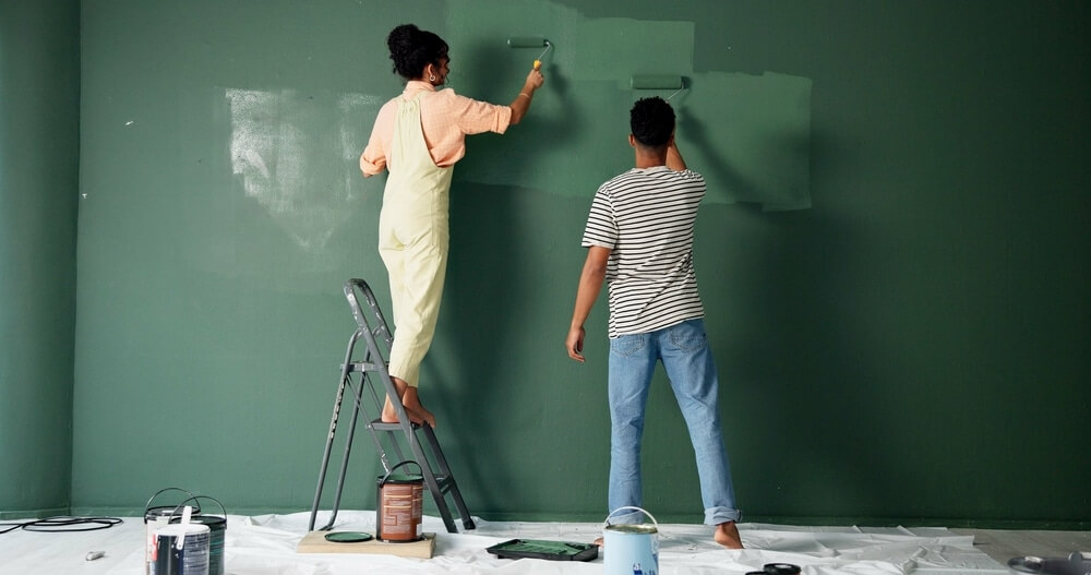 Two people painting a green wall together; one stands on a ladder, and the other works from the floor. Paint cans and tools are spread on a protective sheet below.