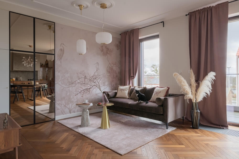 A stylish living room with a beautifully refinished herringbone-pattern parquet wood floor. The room features a plush velvet sofa with decorative pillows, soft pink curtains, and pampas grass in a vase for a cozy, modern aesthetic. A large mirror and elegant lighting fixtures complement the space, along with artistic wall decor.