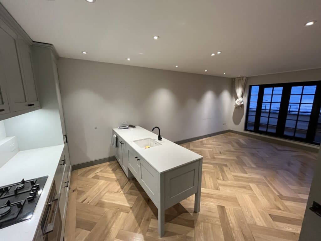 A modern kitchen and living area with a beautifully refinished herringbone-patterned wooden floor, featuring sleek cabinetry and large glass doors.