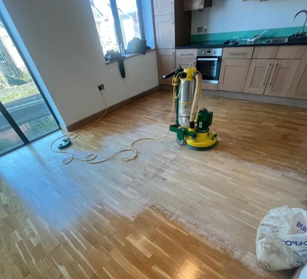 Partially sanded hardwood flooring in a combined kitchen-living area, with a professional floor sanding machine in use, illustrating the process of Floor Sanding for Hardwood Floors.