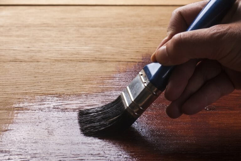 Close-up of a hand using a paintbrush to apply a wood finish on a wooden floor, showcasing the difference between the finished and unfinished sections.