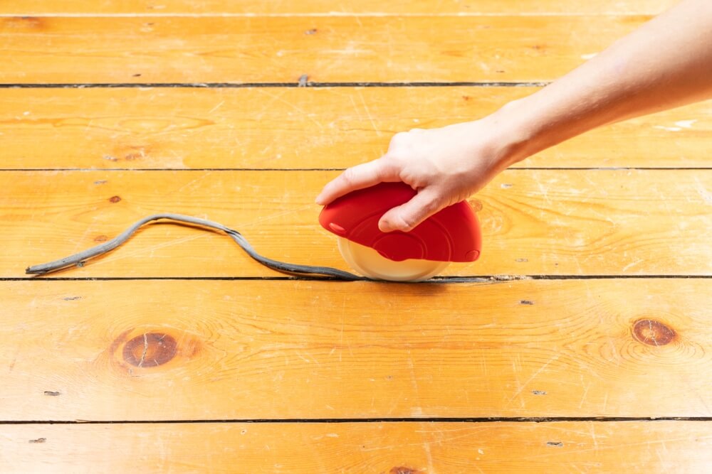 A hand using a red tool to press a rope into the gaps between pine floorboards. The floor has a warm, orange-brown finish, and the process indicates a method of gap filling.
