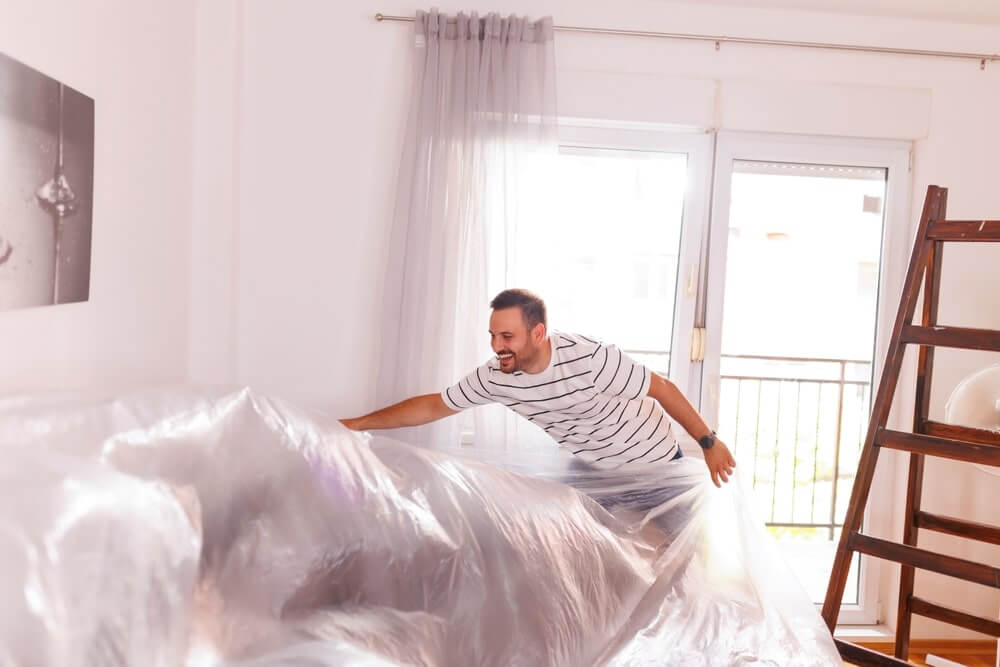A cheerful man prepares a room for floor sanding and refinishing, covering furniture with a plastic sheet. The room features bright natural light from a large glass door and window, with a wooden ladder leaning against the wall. The floor is an oak hardwood surface, ready for transformation.