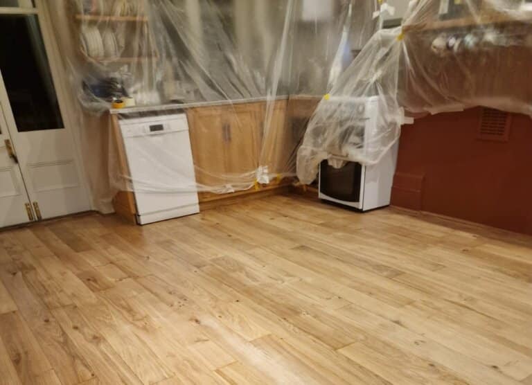 A kitchen prepared for floor sanding and refinishing, with cabinets and appliances covered in protective plastic sheeting. The newly sanded wooden floor is light-colored and clean, showcasing a smooth finish. The floor sanding and refinishing are being completed by Mr Sander®.