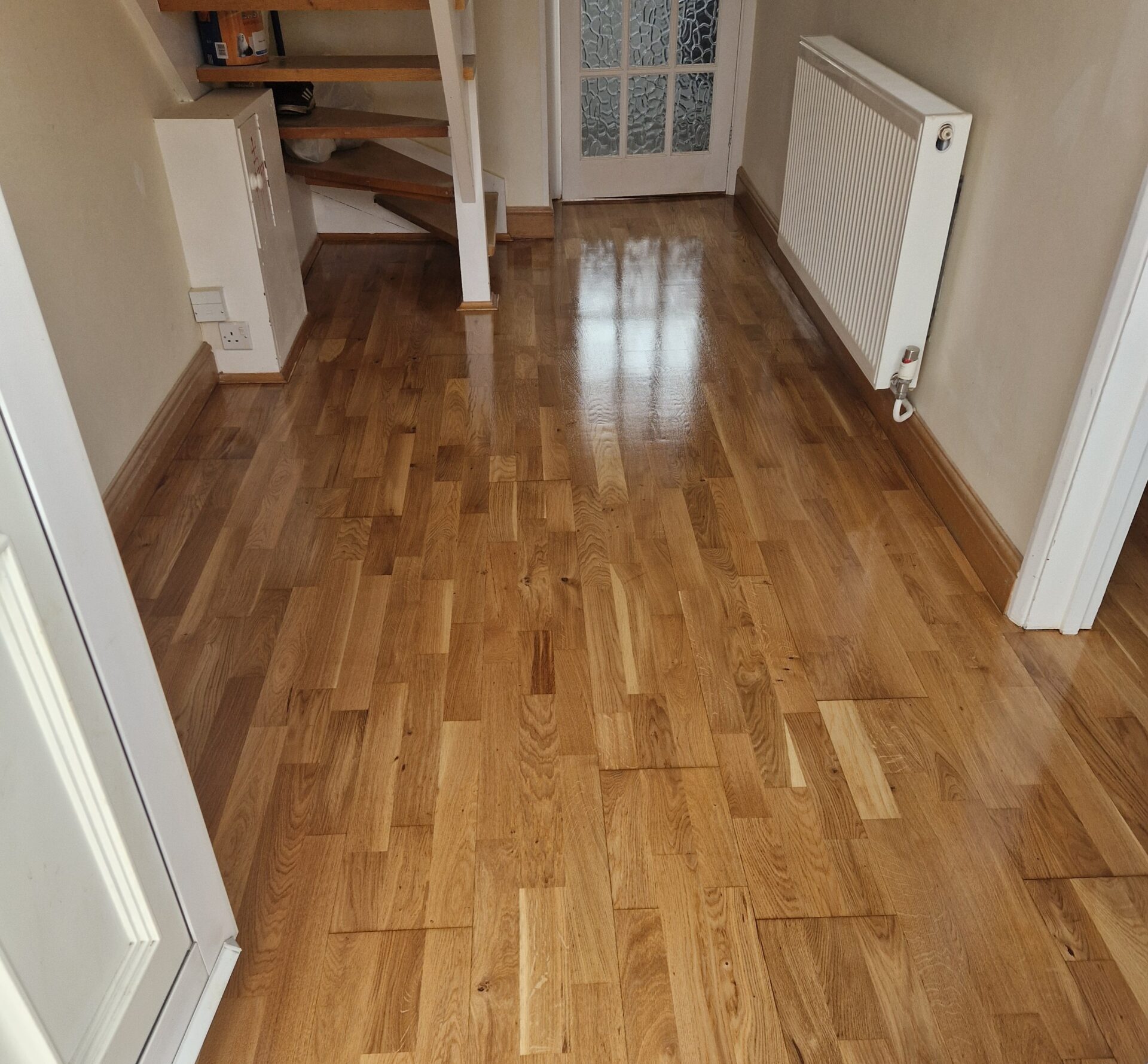A shiny oak wood floor in a hallway with a staircase in the background.
