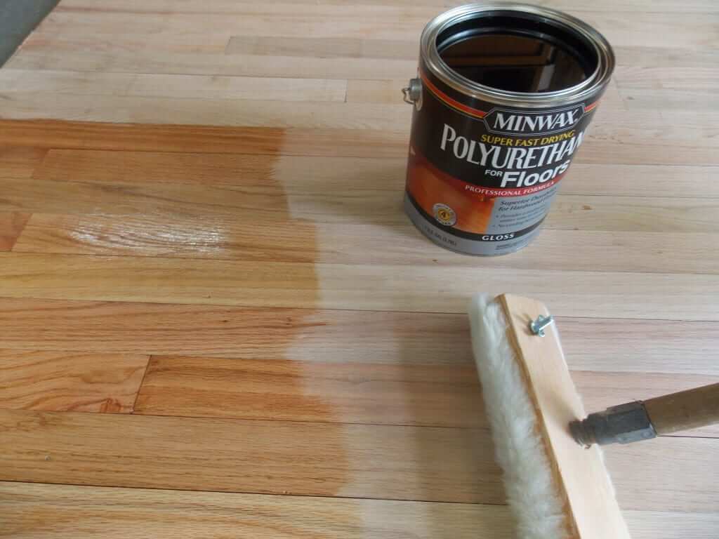A can of Minwax polyurethane for floors sits on a wooden floor as a finish is being applied. A brush applicator is used to spread the polyurethane, with a clear difference between the treated and untreated sections of the floor, highlighting the glossy protective coating being applied.