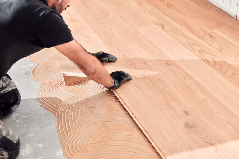 Professional fitter lays engineered oak plank over adhesive swirl during floor installation for smart homes.
