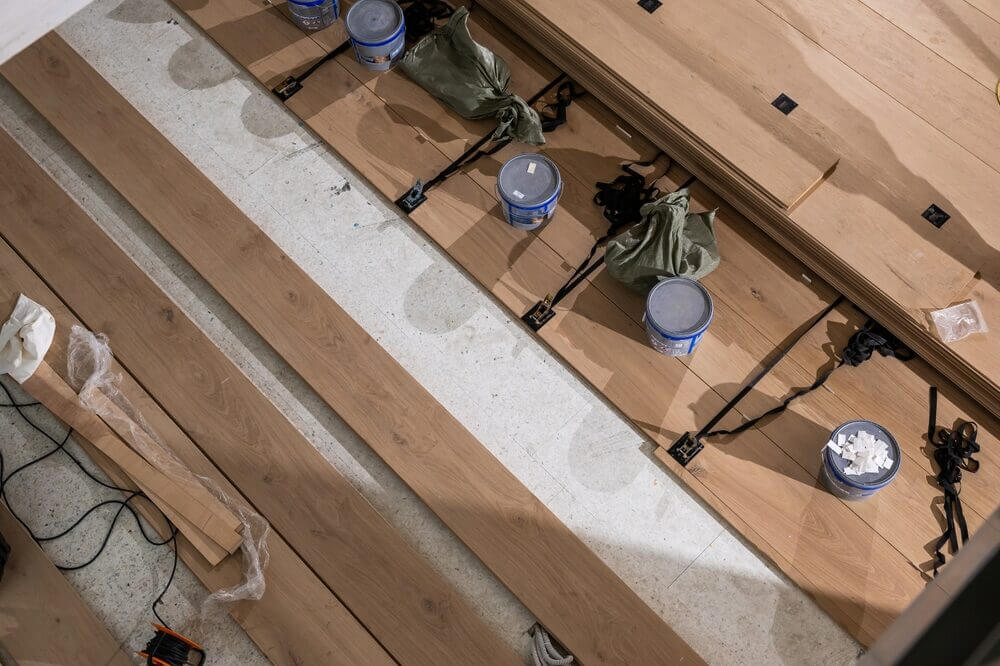 A top-down view of wide wooden planks being glued and clamped in place on a prepared subfloor during a professional installation process.