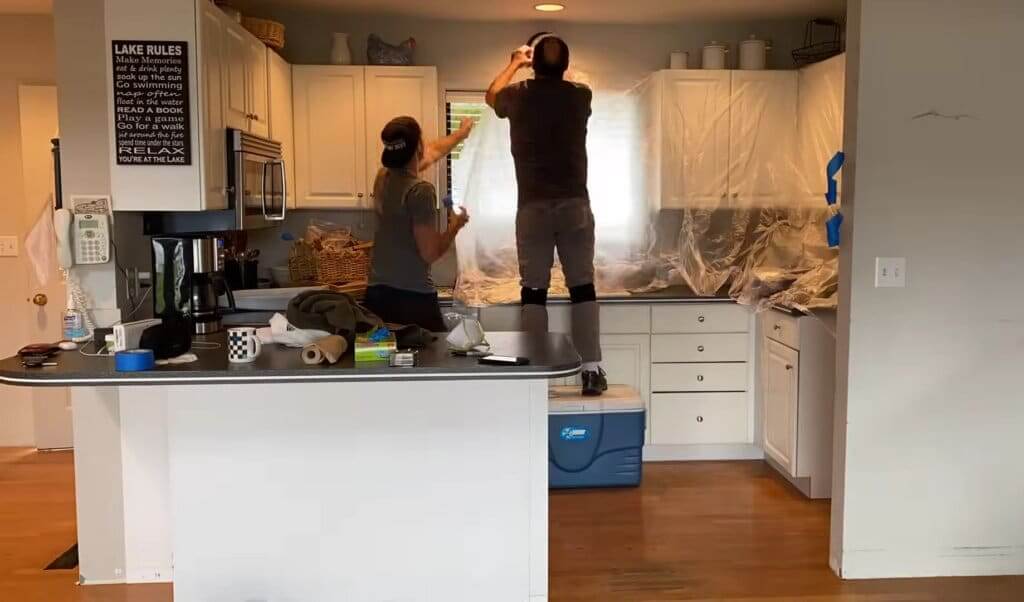 Two workers prepare a kitchen for floor sanding and refinishing by covering the countertops, cabinets, and appliances with protective plastic sheets. One worker stands on a cooler, adjusting the plastic near a window, while the other assists. The kitchen has white cabinets, and various tools and materials are placed on the countertop.