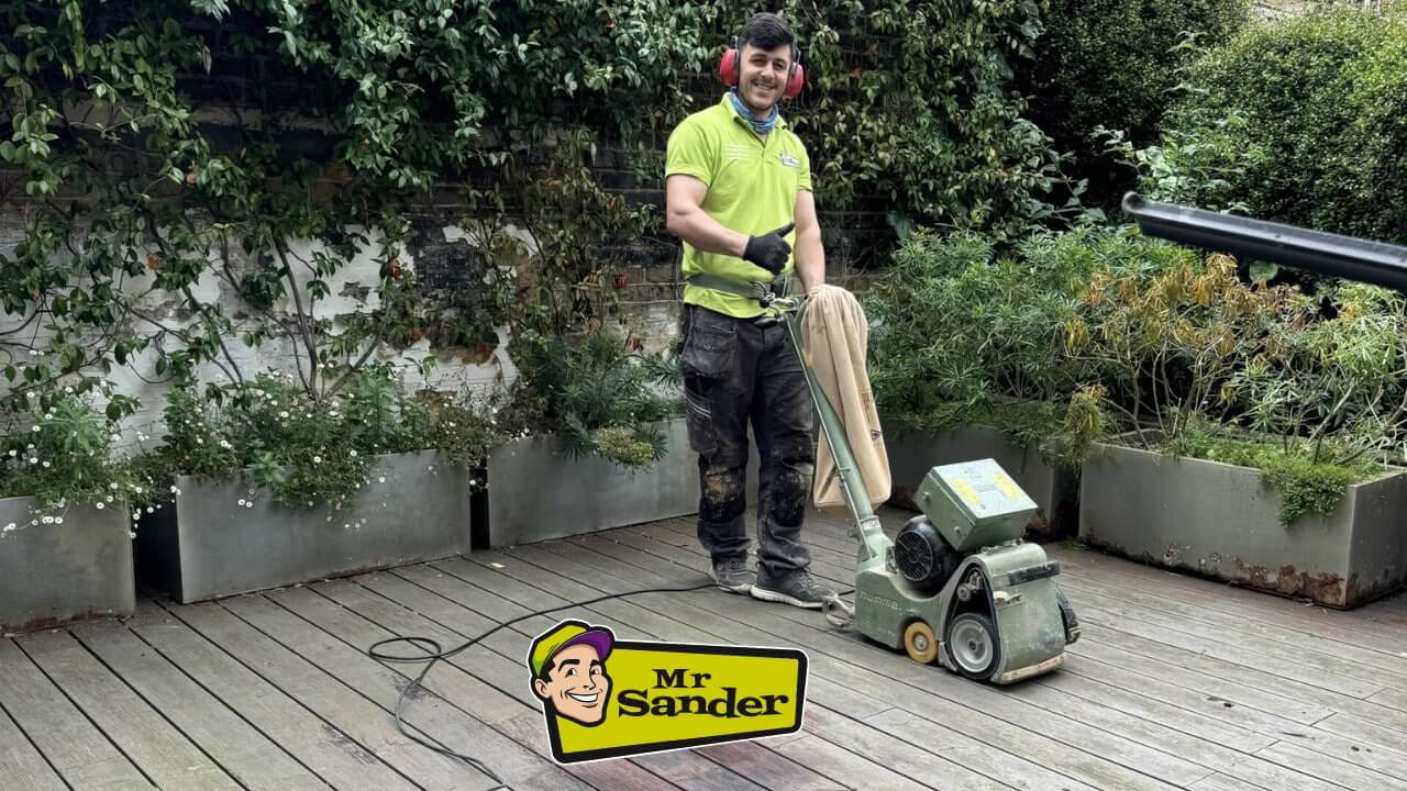 A man from Mr. Sander sanding a wooden deck with a Hummel Lagler sanding machine in a garden setting. The man is wearing a green shirt, protective ear muffs, and gloves, giving a thumbs-up.