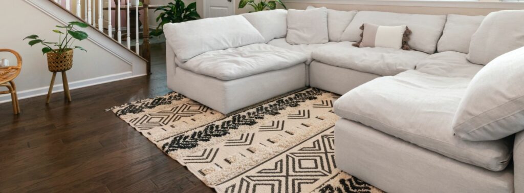 A stylish living room in Deptford, UK, featuring beautifully refinished oak hardwood floors by Mr. Sander. The room includes a cozy white sectional sofa and a patterned area rug, showcasing the contrast and elegance of the dark, polished wooden floor. Plants and a staircase add to the inviting and well-maintained atmosphere, highlighting the professional floor sanding and refinishing services provided by Mr Sander®