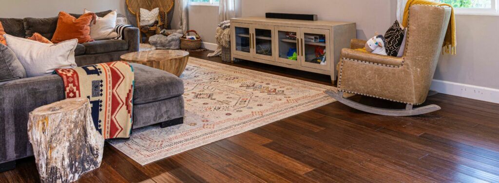 A cozy living room in Tonbridge, UK, featuring beautifully sanded and refinished dark oak wood floors. The space includes a gray sectional sofa with colorful cushions, a rustic wooden coffee table, a rocking chair, and a patterned area rug, showcasing the expert floor sanding and refinishing work by Mr Sander®.