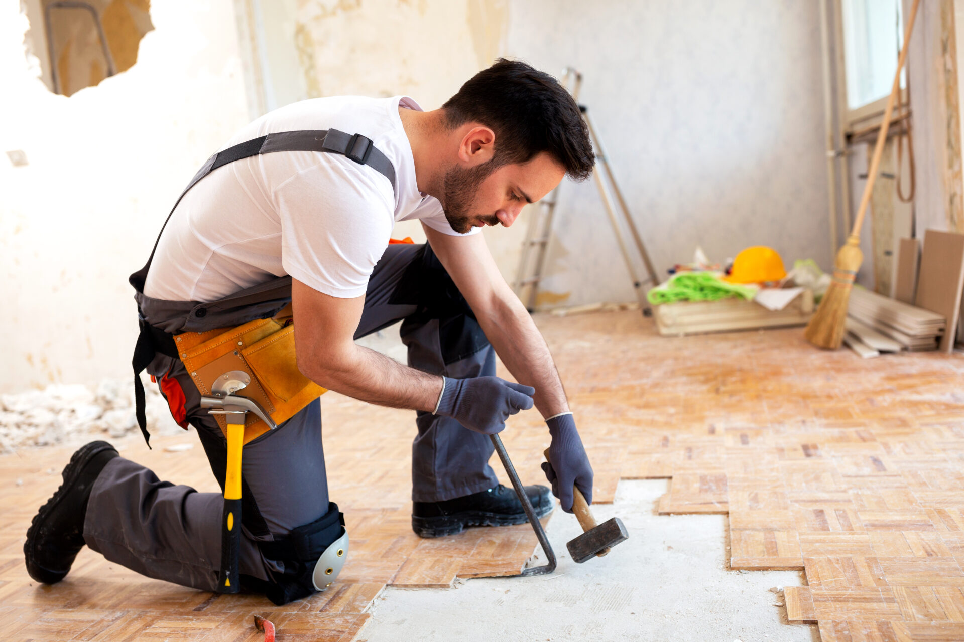 A skilled tradesman kneeling while removing damaged parquet tiles with a crowbar and hammer, preparing the floor for sanding and repair.