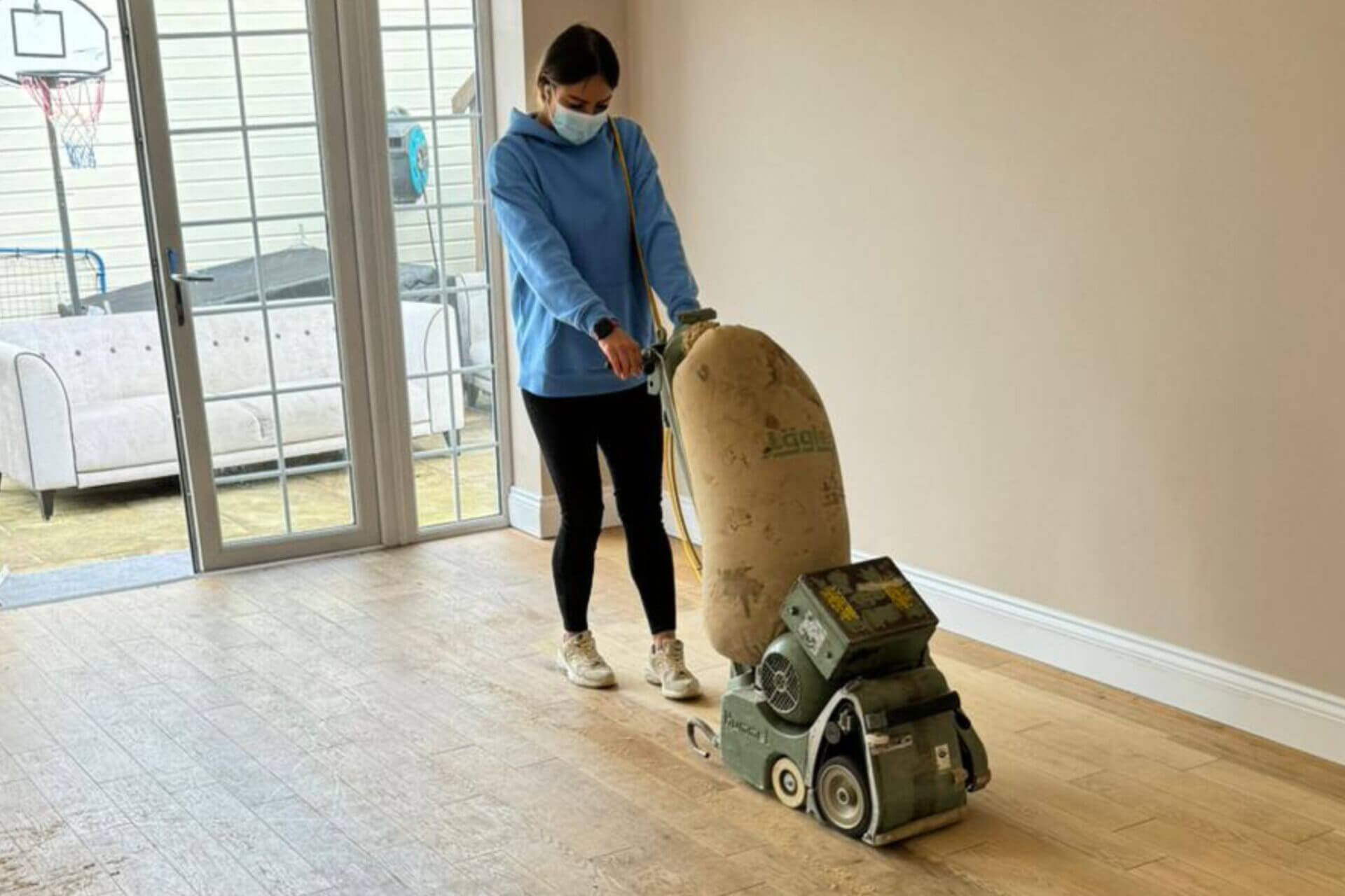 A female technician wearing a face mask carefully operates a floor sanding machine on engineered wood planks near large patio doors.