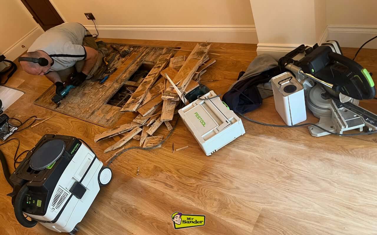A tradesperson working on a hardwood floor section, removing damaged boards with power tools and surrounded by replacement planks.