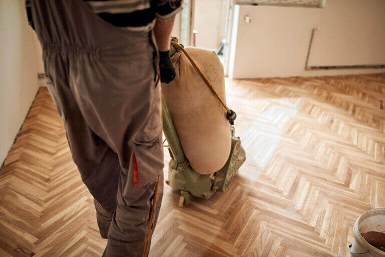 A worker sanding a parquet wood floor with a drum sander, showcasing the process of Sanding Different Types of Wood Floors.