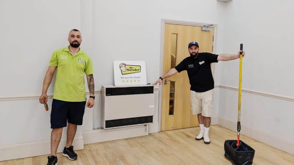 Two workers from Mr. Sander® standing in a room with freshly sanded and refinished wooden floors. The worker on the left is wearing a green shirt, holding a tool, while the worker on the right is wearing a black shirt, holding a mop. They are standing next to a sign that reads "We give Mr. Sander® 5 stars" and a wooden door. The room is clean and bright, showcasing the quality of their work.