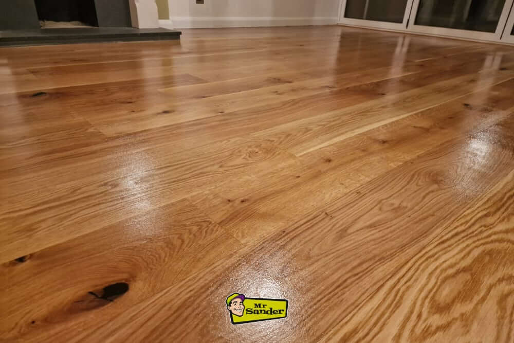A close-up view of a beautifully restored oak wooden floor with a glossy finish, reflecting the light in a living room setting. A fireplace and large glass doors are visible in the background.