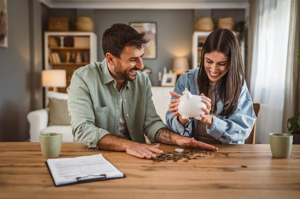 A smiling couple sits at a wooden table, counting coins from a piggy bank, symbolizing savings. The woman pours coins onto the table while the man organizes them. In the background, a cozy living room with warm lighting and wooden shelves creates a welcoming atmosphere. The natural grain of the wooden table complements the couple's happy expressions as they review a clipboard with paperwork and two mugs beside them.