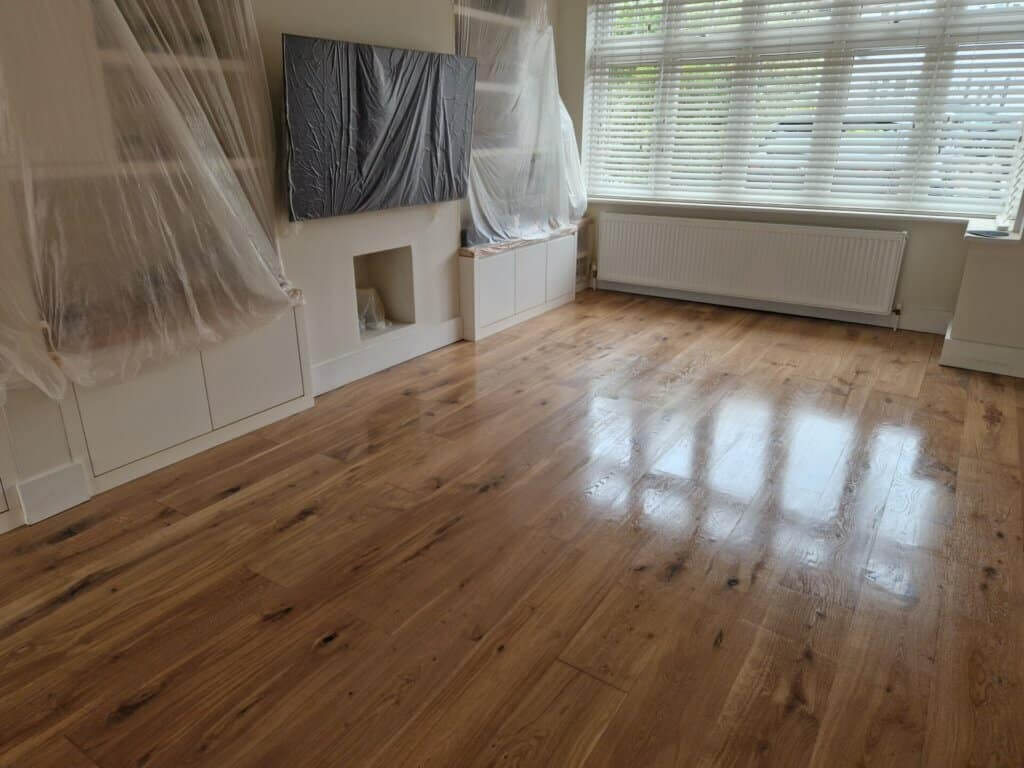 A bright living room featuring a newly sanded and refinished oak wood floor with a glossy, polished finish. The natural grain of the wide oak planks adds warmth and character to the space. Furniture and wall fixtures are covered with protective plastic sheets, emphasizing the refinishing work in progress. A large window with blinds allows natural light to reflect off the shiny floor surface. The overall look is clean and modern.