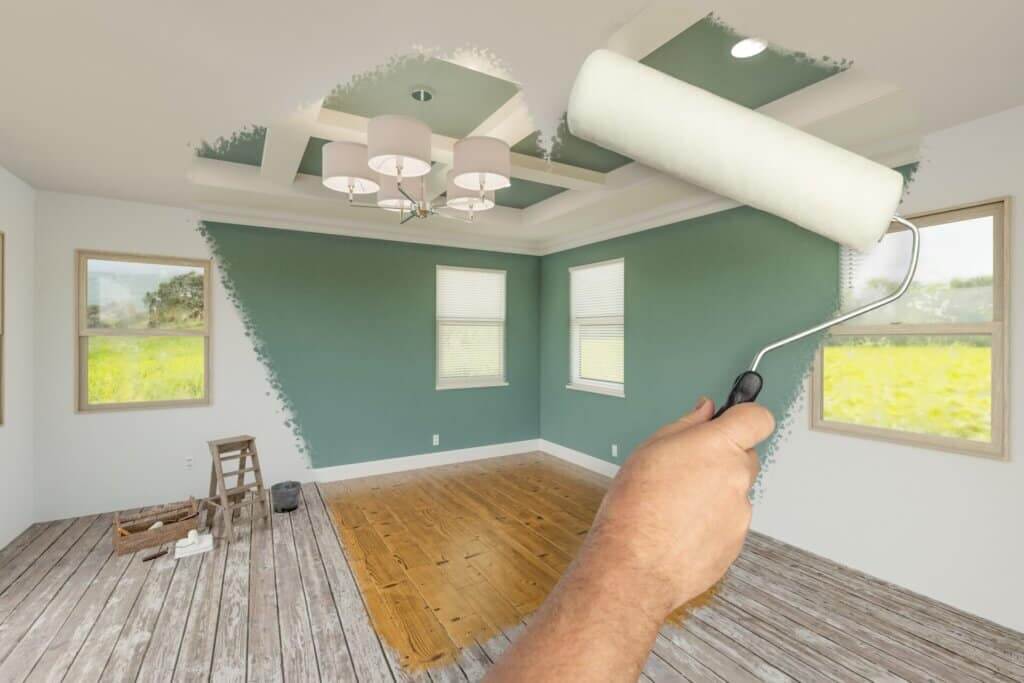 A hand holding a paint roller in the foreground, painting a green accent wall in a room with wooden floors in progress. The unfinished wood floor transitions to a polished, warm-toned finish in the image.