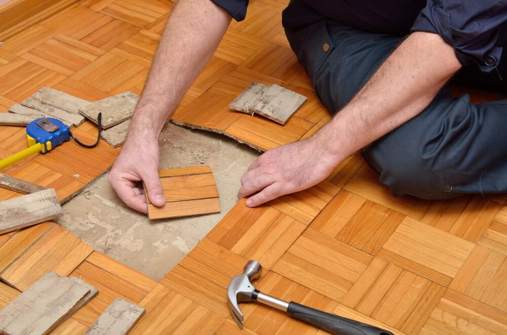A person is seen repairing a wooden parquet floor. The individual is placing a replacement wooden tile into a damaged section of the floor. Various tools, including a tape measure, hammer, and a pry bar, are scattered nearby, indicating a work in progress.