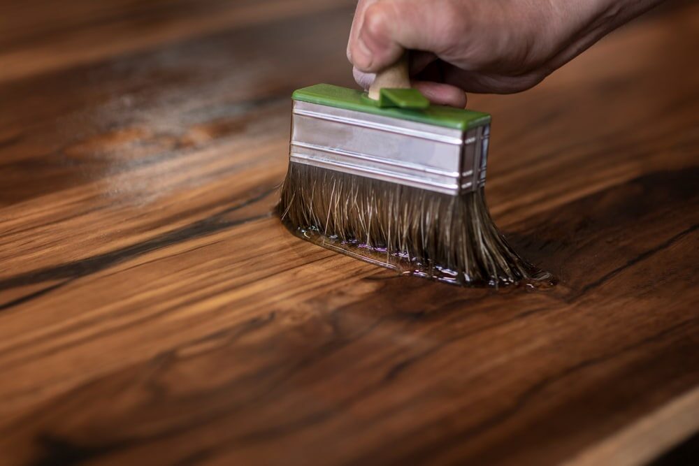 Close-up of a hand applying stain with a brush on a wooden surface.