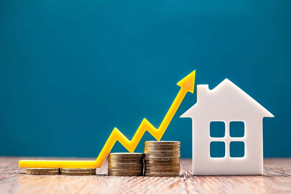 A stack of coins gradually increasing in height is placed next to a small house model. A yellow arrow curves upwards, symbolizing growth in value or property prices. The background is a blue gradient, emphasizing the concept of increasing home market value.