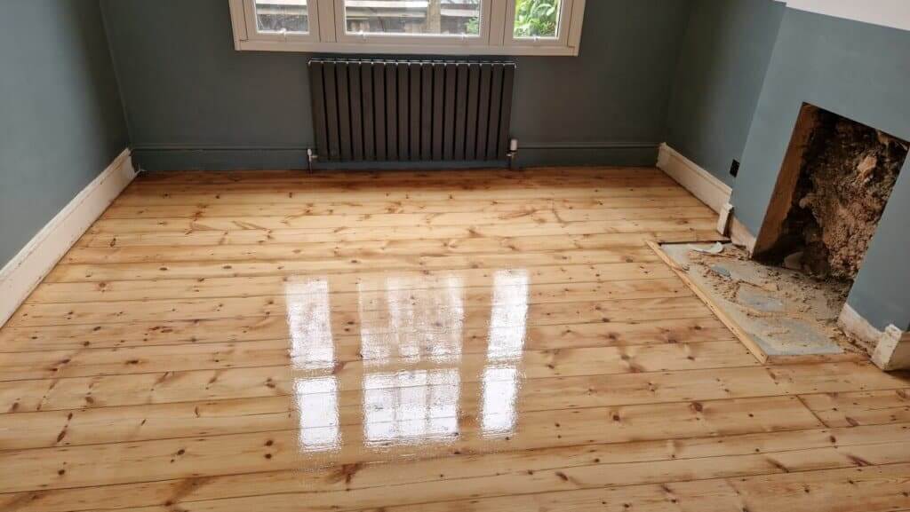 A room with freshly restored pine wood flooring that has a glossy finish. The floor reflects light from the windows, highlighting the natural grain and smooth surface. The room has dark grey walls, a radiator under the window, and an unfinished fireplace area, suggesting ongoing renovations.