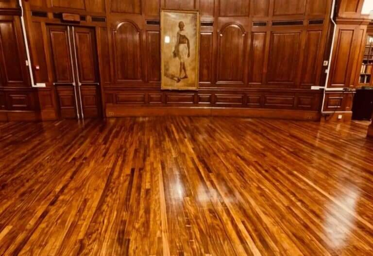 A beautifully restored wooden floor with a glossy finish, set within a grand room with dark wooden wall paneling and a framed painting on the back wall. The floor appears to have been recently sanded and refinished, showcasing the natural grain and sheen of the wood.