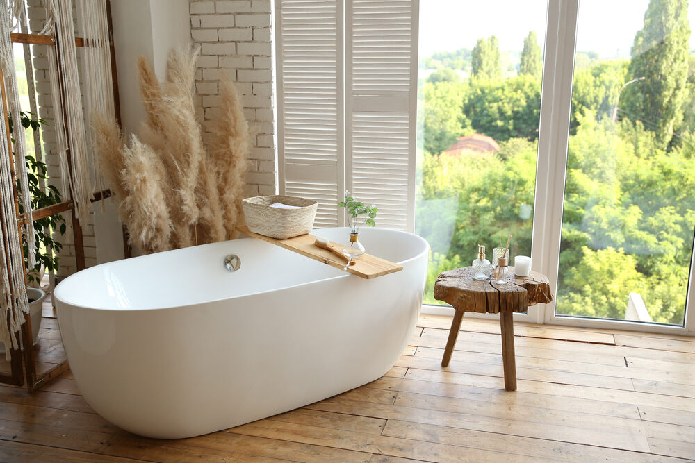 A freestanding bathtub on a rustic wooden floor in a bright, nature-inspired bathroom, illustrating the benefits of ‘Floor Sanding for Bathrooms.