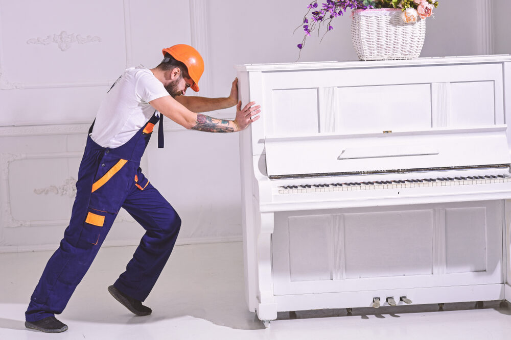 A professional mover in a helmet pushing a white upright piano in a bright, ornate room.