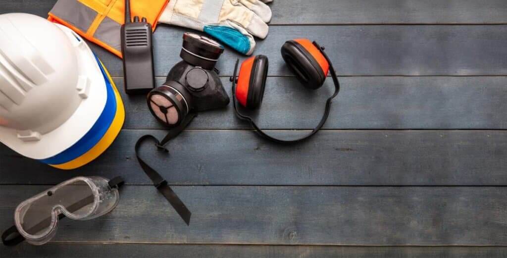 A collection of safety equipment laid out on a wooden surface, including a hard hat, safety goggles, gloves, a respirator mask, ear protection, and a high-visibility vest. These items are essential for ensuring safety during tasks like floor sanding and refinishing.