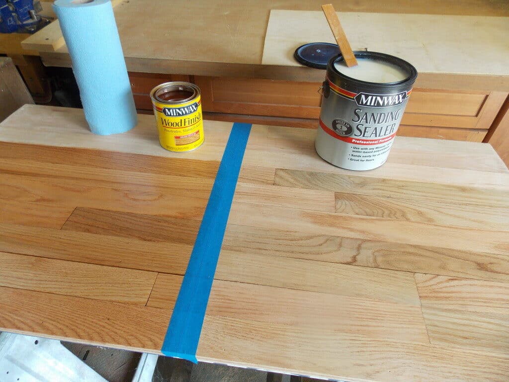 Half-stained wooden plank with Minwax products and a blue painter's tape dividing line.