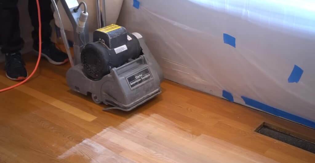 A floor sanding machine in action, being used to refinish a hardwood floor. The machine is removing the top layer of the floor, revealing a lighter, unfinished wood beneath. Protective plastic sheets are taped to the wall, safeguarding the area from dust during the sanding process.
