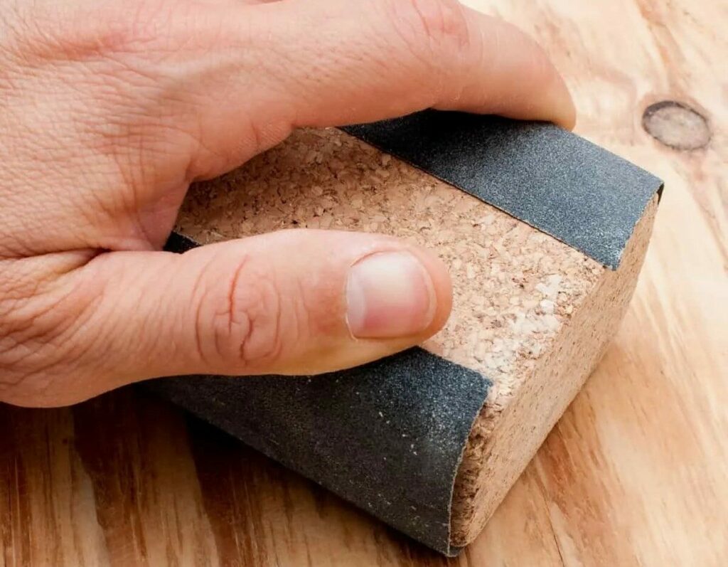 A person wearing gloves carefully sands a piece of wood with a cork block and sandpaper, smoothing out the surface.