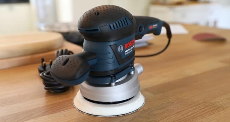 A Bosch random orbit sander placed on a wooden surface, with a power cord coiled beside it.