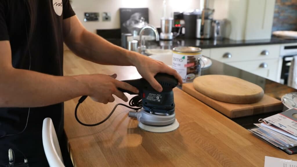 "Person using an orbital sander on a wooden kitchen countertop with kitchen appliances in the background.